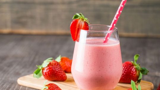 Glass of fresh strawberry milkshake, smoothie and fresh strawberries on pink, white and wooden background. Healthy food and drink concept.